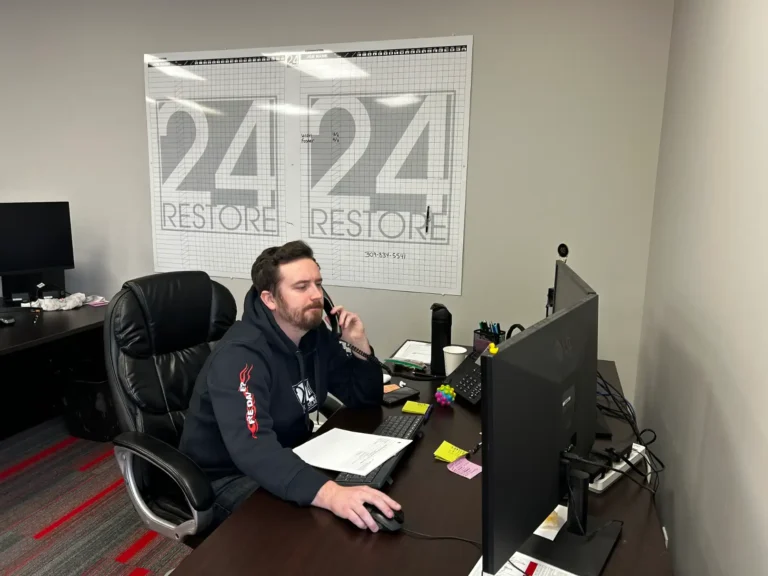 A man sits at a desk in an office, using a computer and speaking on the phone. Papers, a notepad, and office supplies are on the desk. The wall displays a "24 Restore" sign as he discusses finance options with the internal claims team.