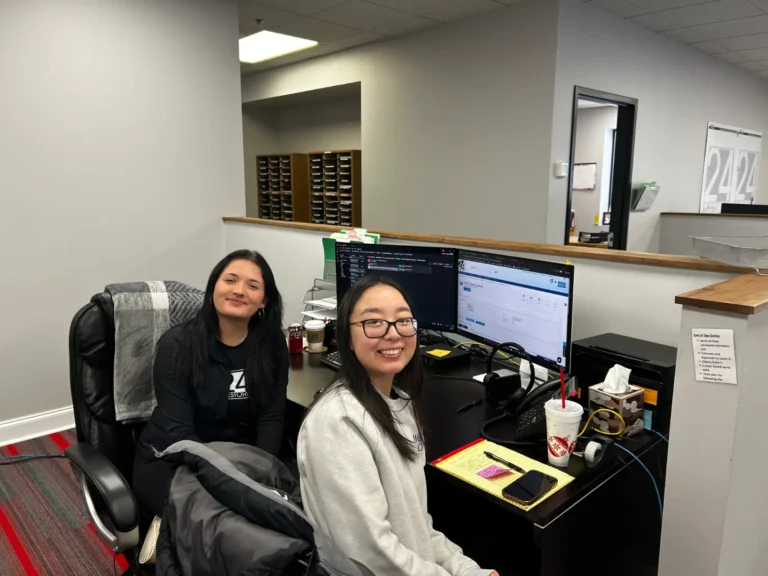 Two people sit at a desk with computers and paperwork in an office space, smiling at the camera. Various office supplies, drinks, and documents related to finance options are visible on the desk.