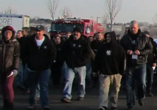A group of people dressed in casual winter clothing walk together outdoors, with a red fire truck and leafless trees visible in the background.